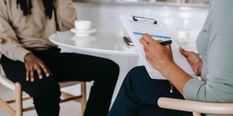 Two people sitting around a white table reviewing contents on a clipboard.|A person sitting behind a glass wall reviewing their computer.